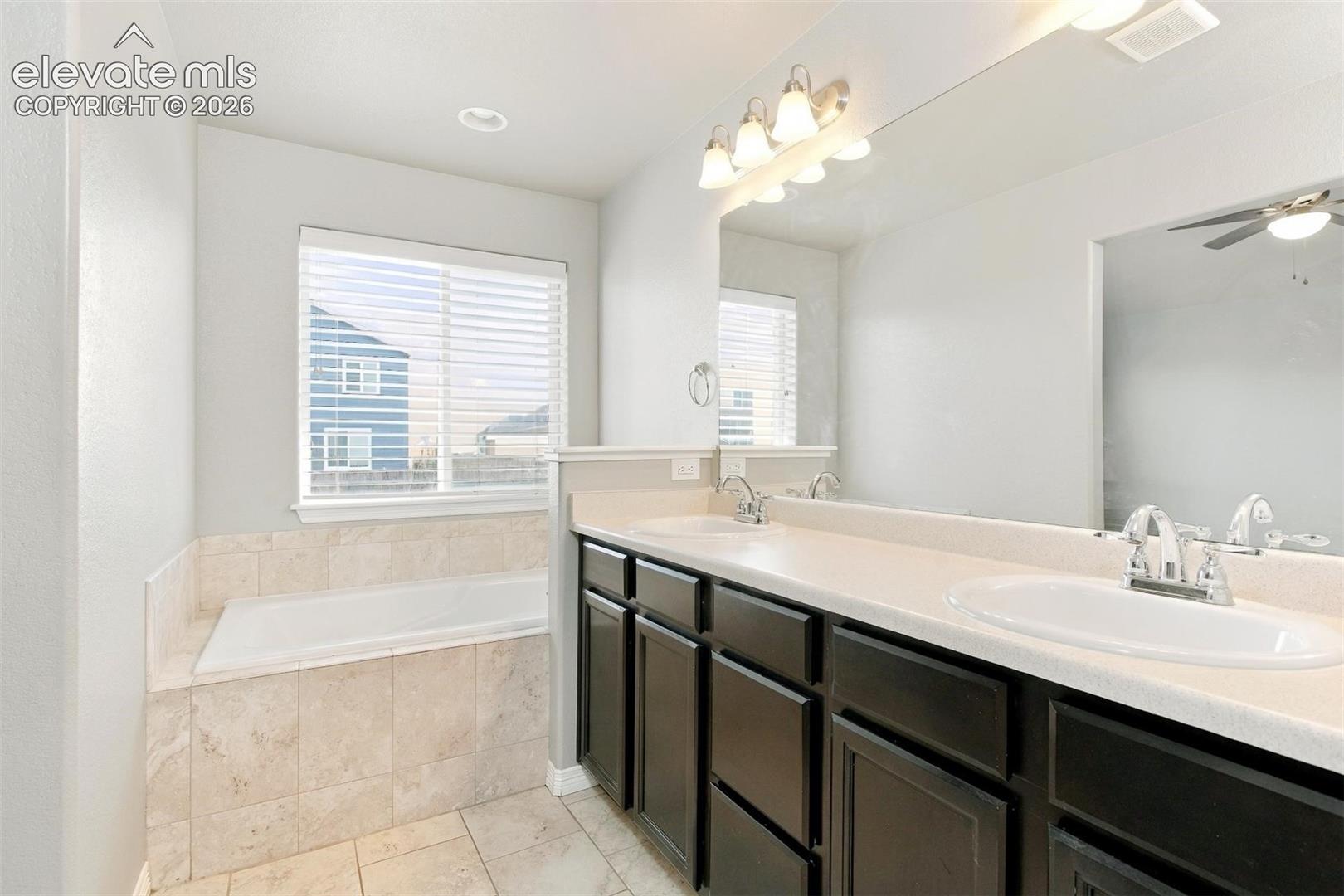 Double vanity featuring dark cabinetry and a solid-surface countertop with dual under-mount sinks, paired with a deep soaking tub featuring tile surround and flooring