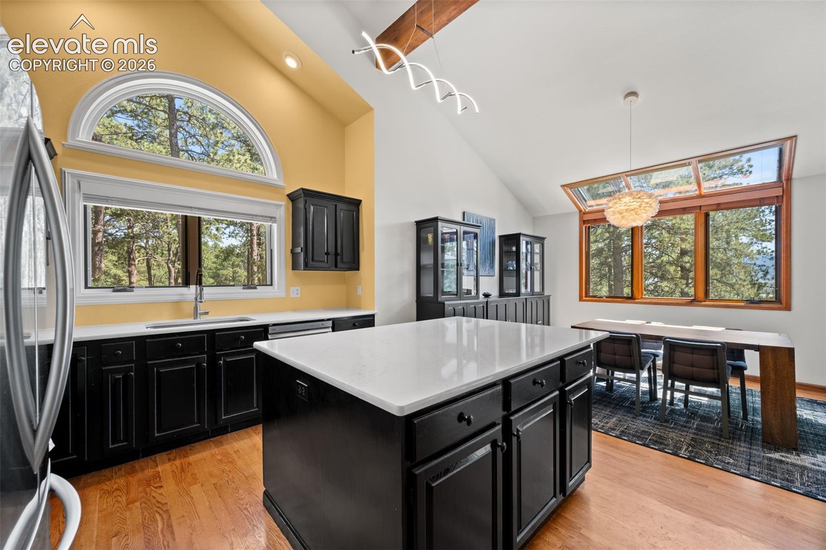 Open-concept kitchen and dining area featuring vaulted ceilings with exposed wood beams, wood-finish flooring, and expansive window walls