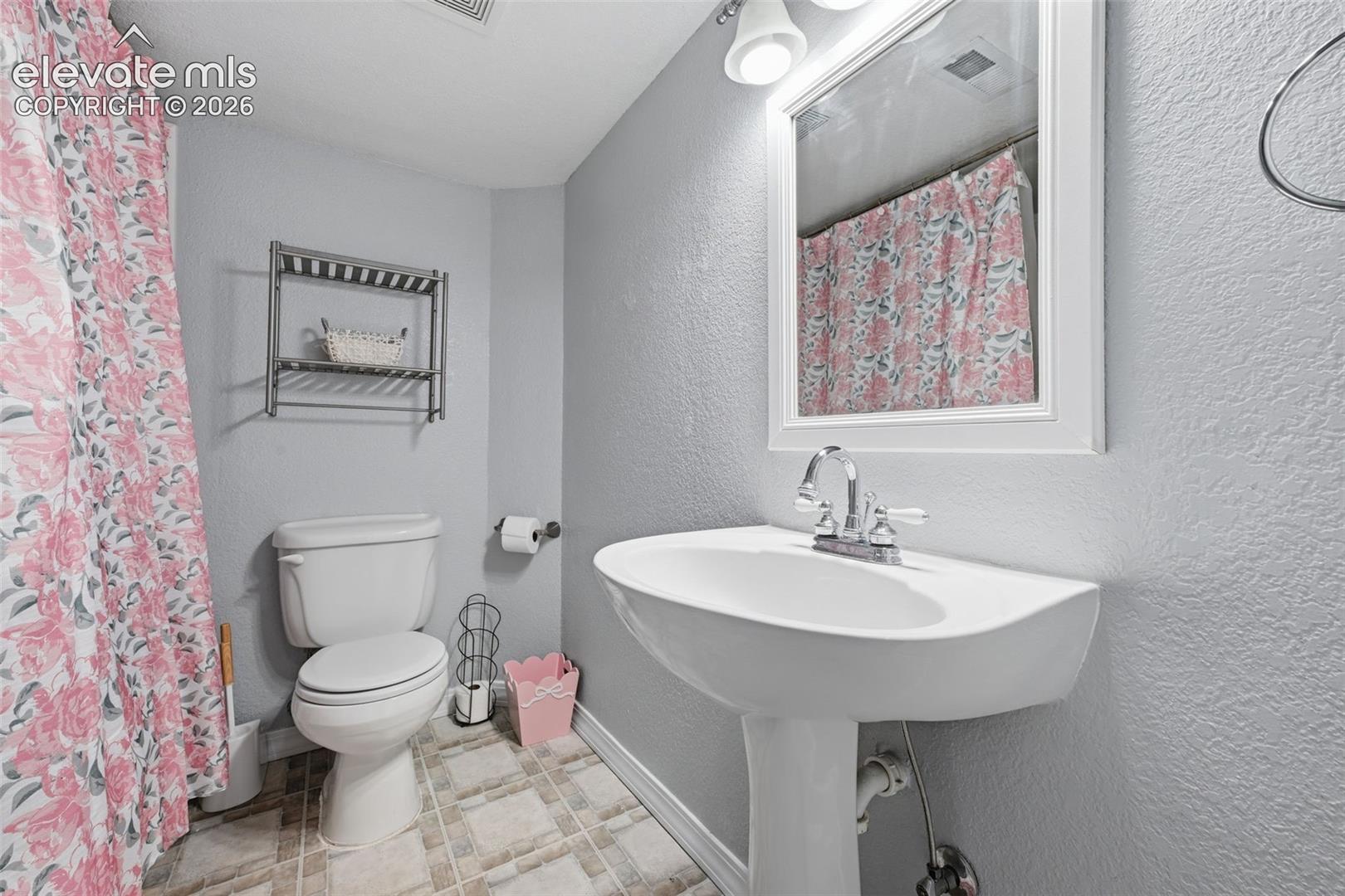 Pedestal sink with chrome faucet, framed vanity mirror, overhead vanity lighting, white commode, and patterned floor tile