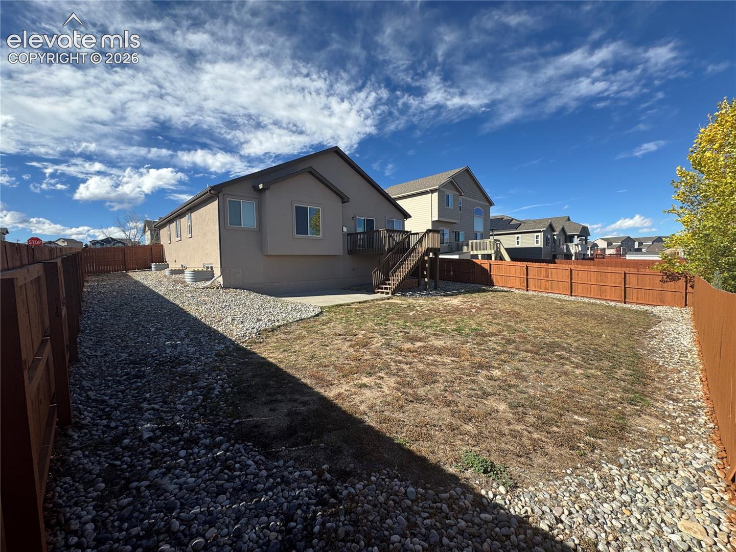 Rear yard featuring a full-perimeter wood privacy fence, stone ground cover, a concrete patio, and an elevated wood deck with stairs