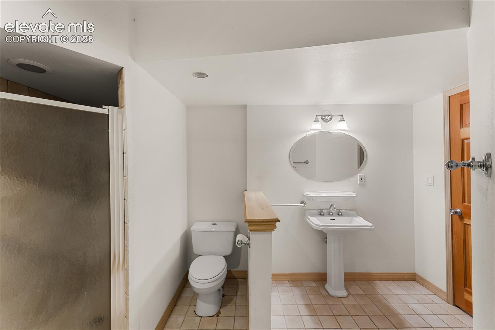 Bathroom featuring a glass-enclosed shower, pedestal sink, oval vanity mirror with sconce lighting, and tile flooring