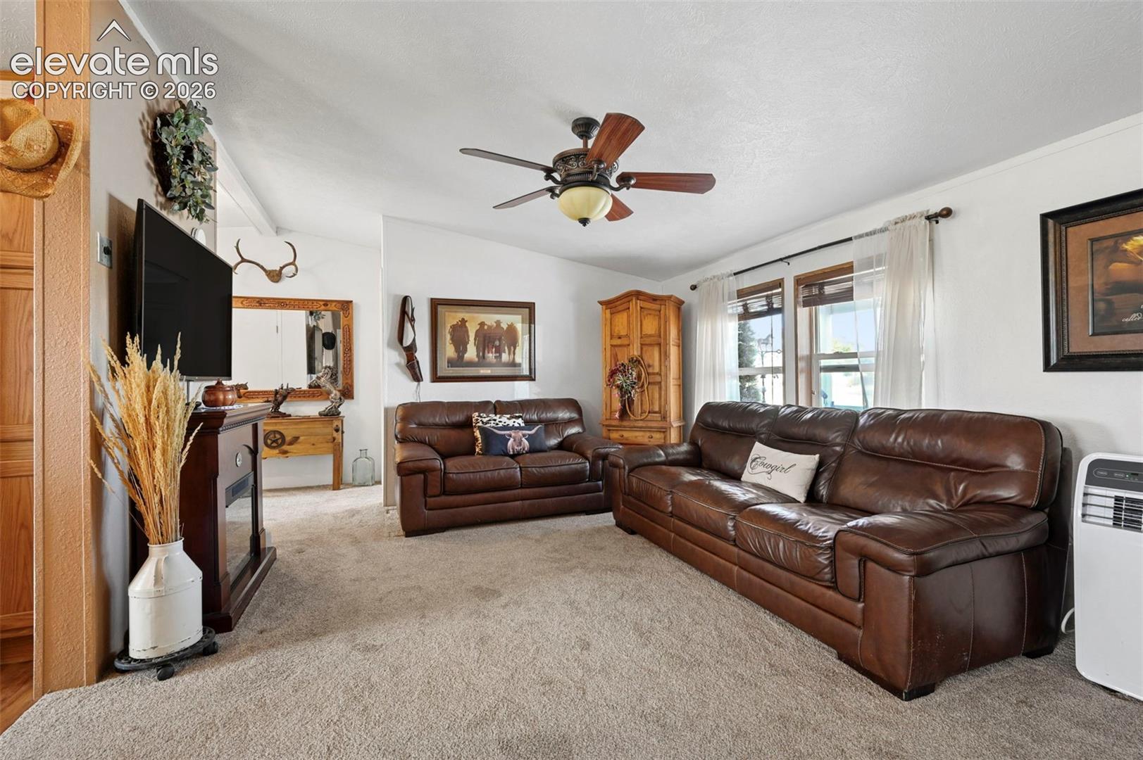 Living area featuring wall-to-wall carpeting, vaulted ceilings, multi-blade ceiling fan with integrated lighting, and dual windows with wood trim