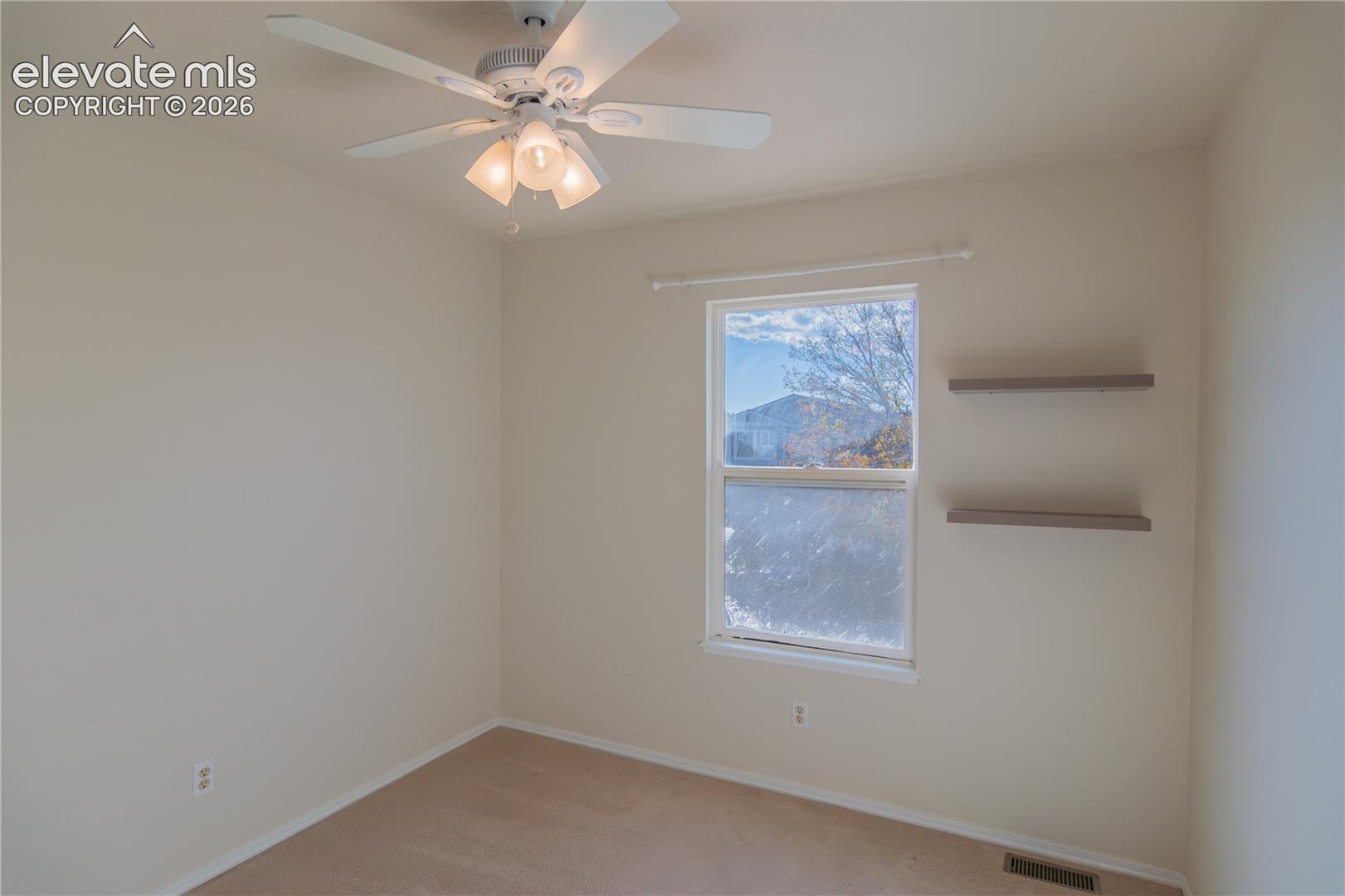 Bright secondary bedroom with a ceiling fan, neutral carpeting, and decorative wall shelving.