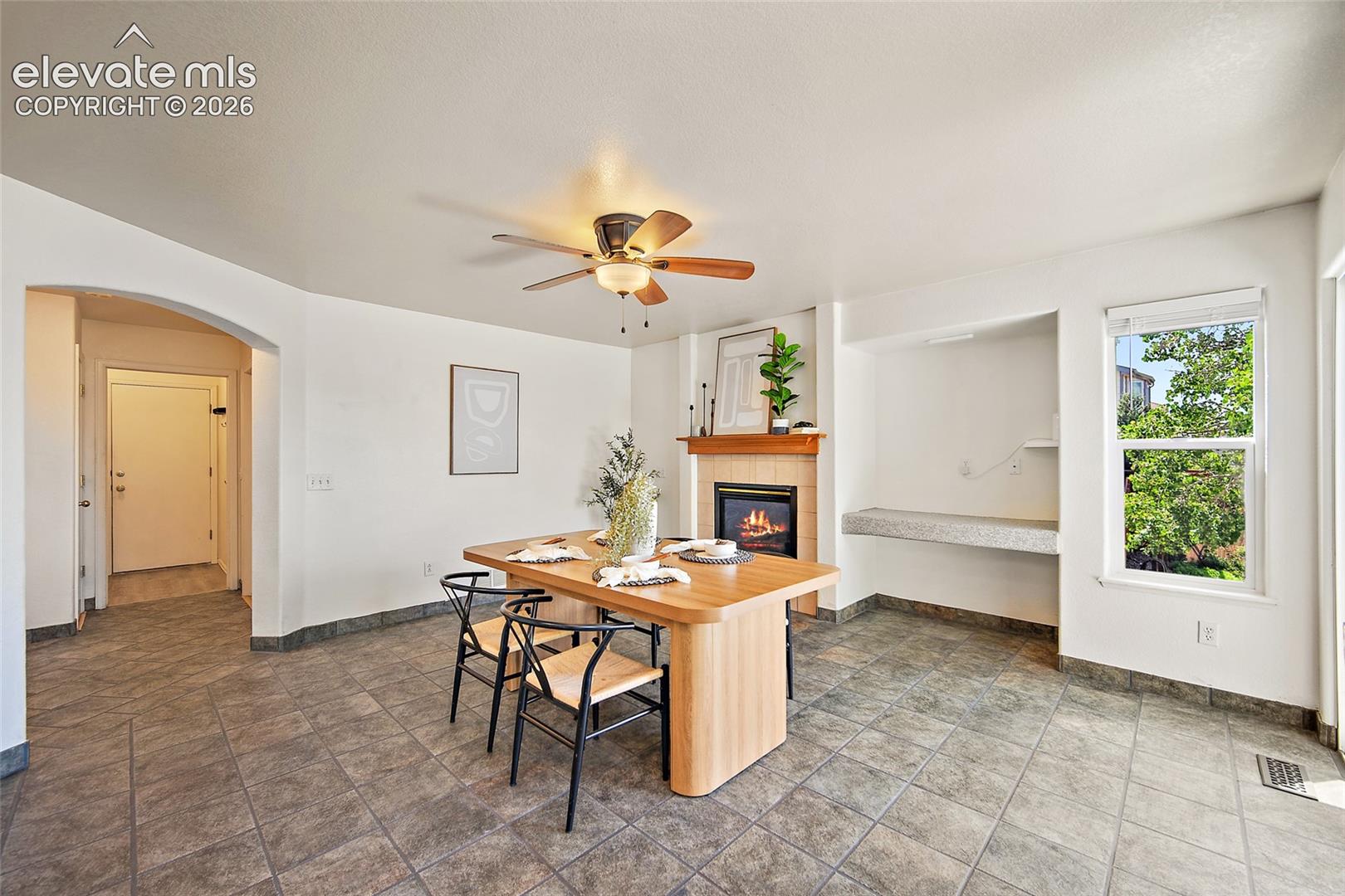 Spacious room featuring tile flooring, a built-in fireplace with a wood mantle, an arched doorway, and a ceiling fan with integrated lighting