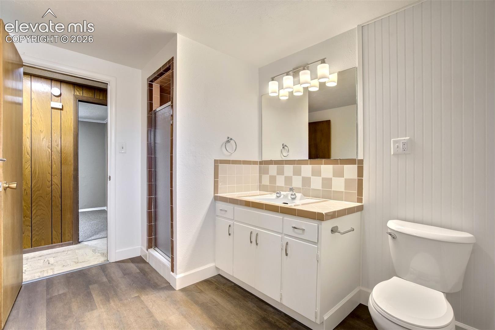 Bathroom featuring a vanity with white cabinetry, tile countertop, beadboard wall paneling, wood-finish flooring, and a glass-enclosed shower