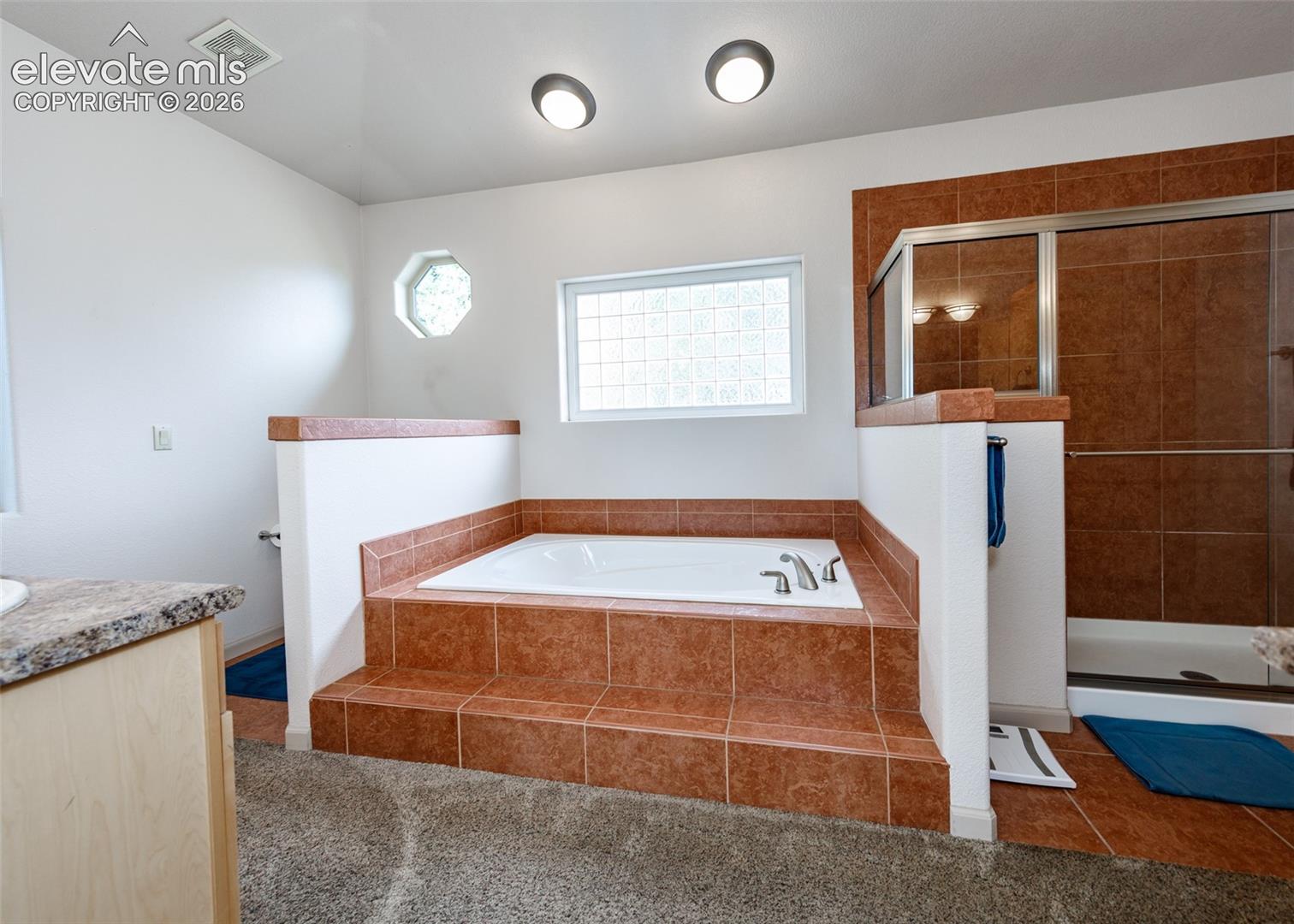 Built-in soaking tub with tiled surround, separate shower enclosure with glass doors, octagonal window, and dual vanities with stone countertop