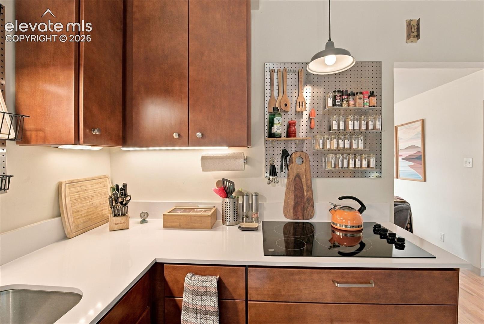 Kitchen featuring rich wood-finish cabinetry, solid surface countertops, a glass cooktop, under-cabinet task lighting, and a metal pegboard wall organizer