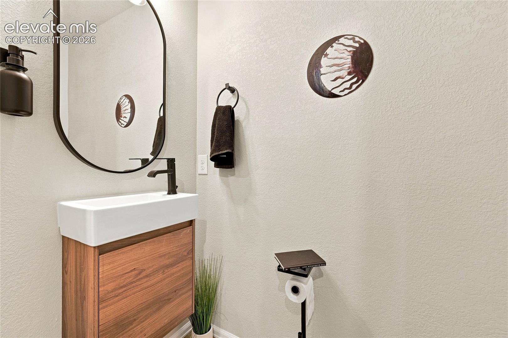 Wall-mounted wood-finish vanity with rectangular white basin, matte black faucet, oval-shaped mirror with dark metal frame, and matching matte black hardware