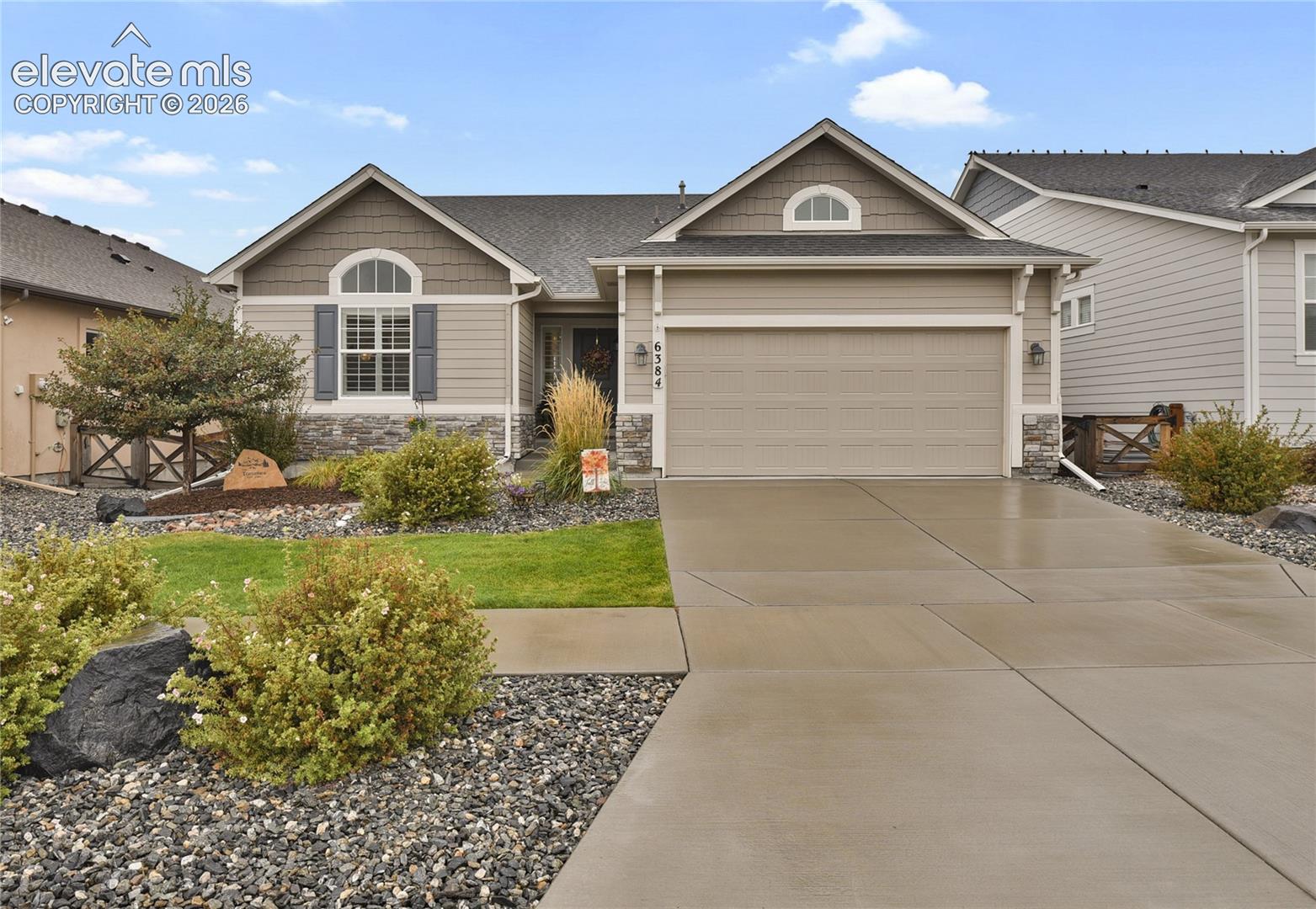 Single-story beige house with a two-car garage, stone accents, and landscaped front yard.