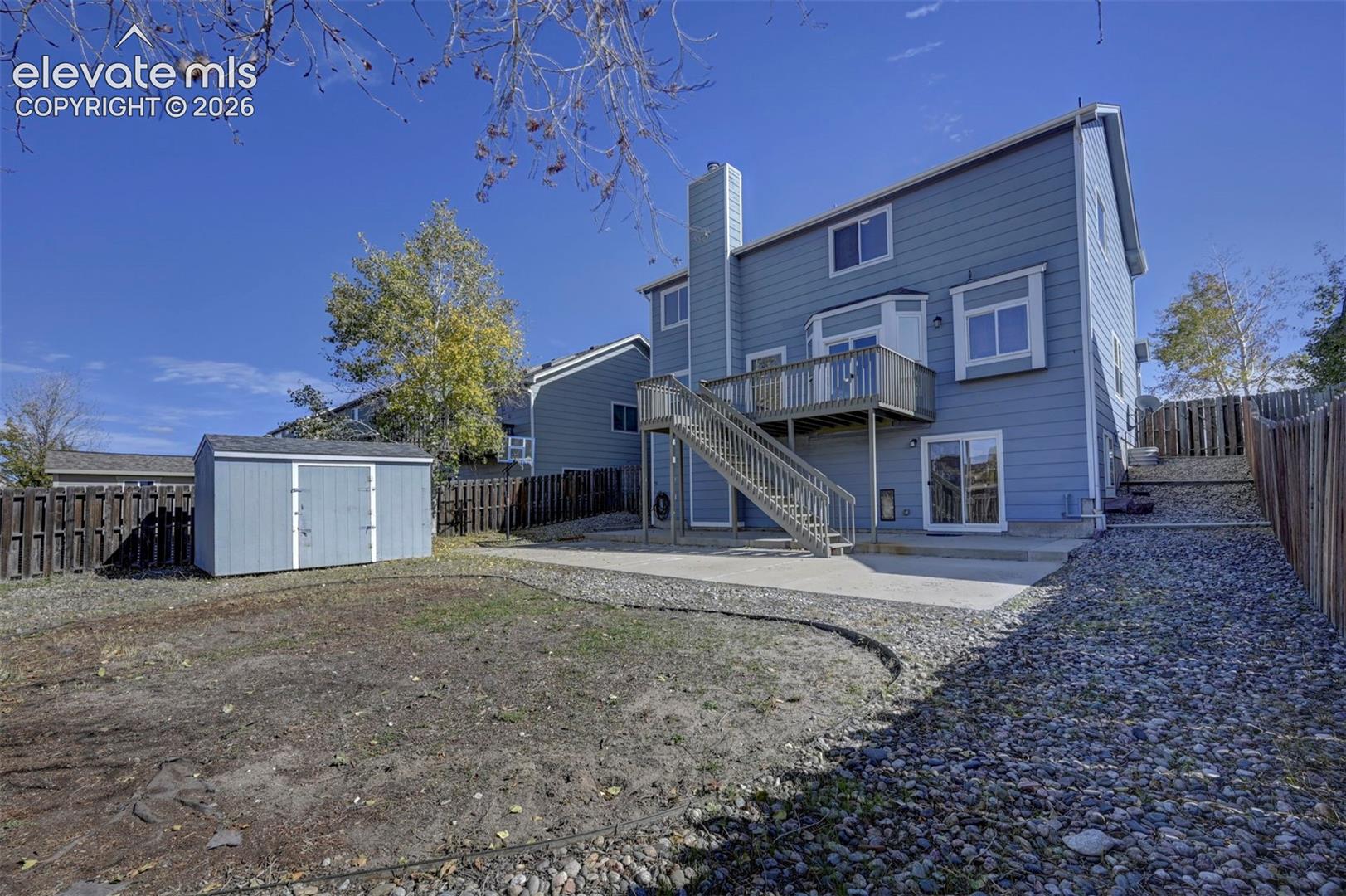 Rear exterior featuring horizontal lap siding, a raised wood deck with staircase, a concrete patio, and a detached storage shed within a fenced yard