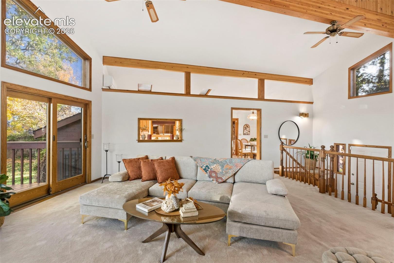 Vaulted ceilings with exposed timber beams, expansive clerestory windows, sliding glass door access to an exterior deck, and a wooden balustrade staircase