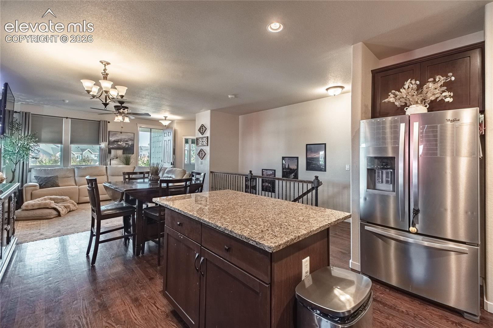 Kitchen island featuring stone countertop and dark wood-finish cabinetry, stainless steel refrigerator, dark wood-finish flooring, recessed lighting, and metal baluster railing