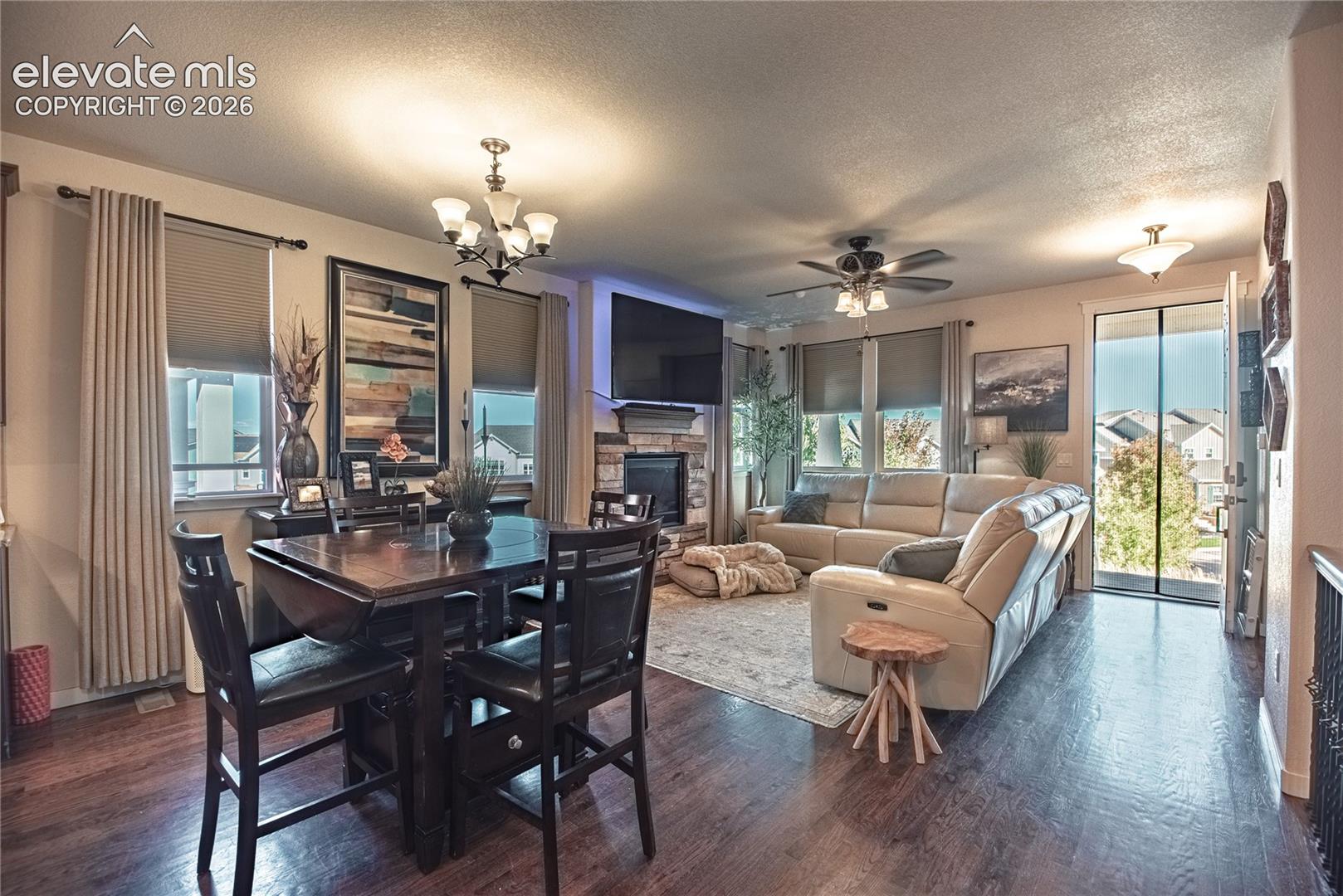 Open concept living area featuring dark wood-finish flooring, stone-surround fireplace, multi-arm chandelier lighting, and a ceiling fan