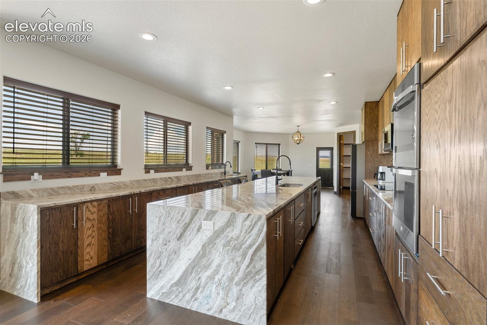 Spacious kitchen featuring a large waterfall island with integrated sink, wood-finish flooring, custom wood cabinetry, stainless steel double ovens, and ample natural light from multiple windows