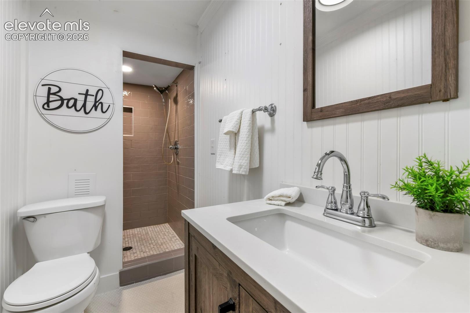 Bathroom featuring beadboard wall paneling, a vanity with a light-toned countertop, an integrated rectangular sink, a framed mirror, and a walk-in shower with tiled surround