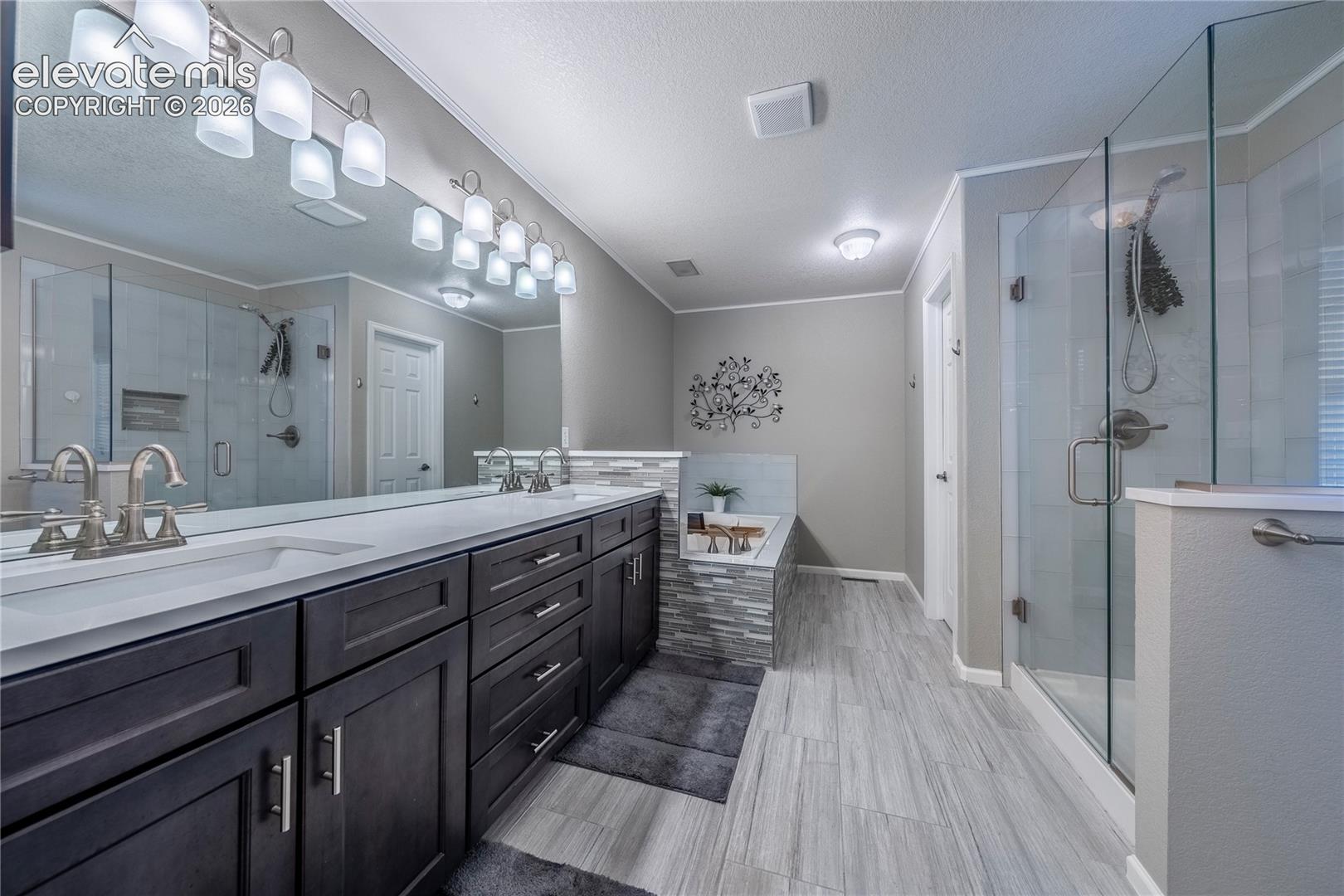 Dark-finish cabinetry with dual undermount sinks, stone-surface countertops, walk-in glass-enclosed shower, soaking tub with tile surround, and wood-finish flooring