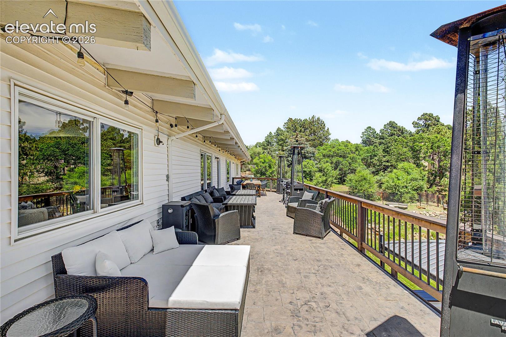 A long, stamped-stone upper deck stretches the full width of the home, strung with café lights and outfitted with multiple seating areas — clearly built for taking in those tree-lined mountain views.