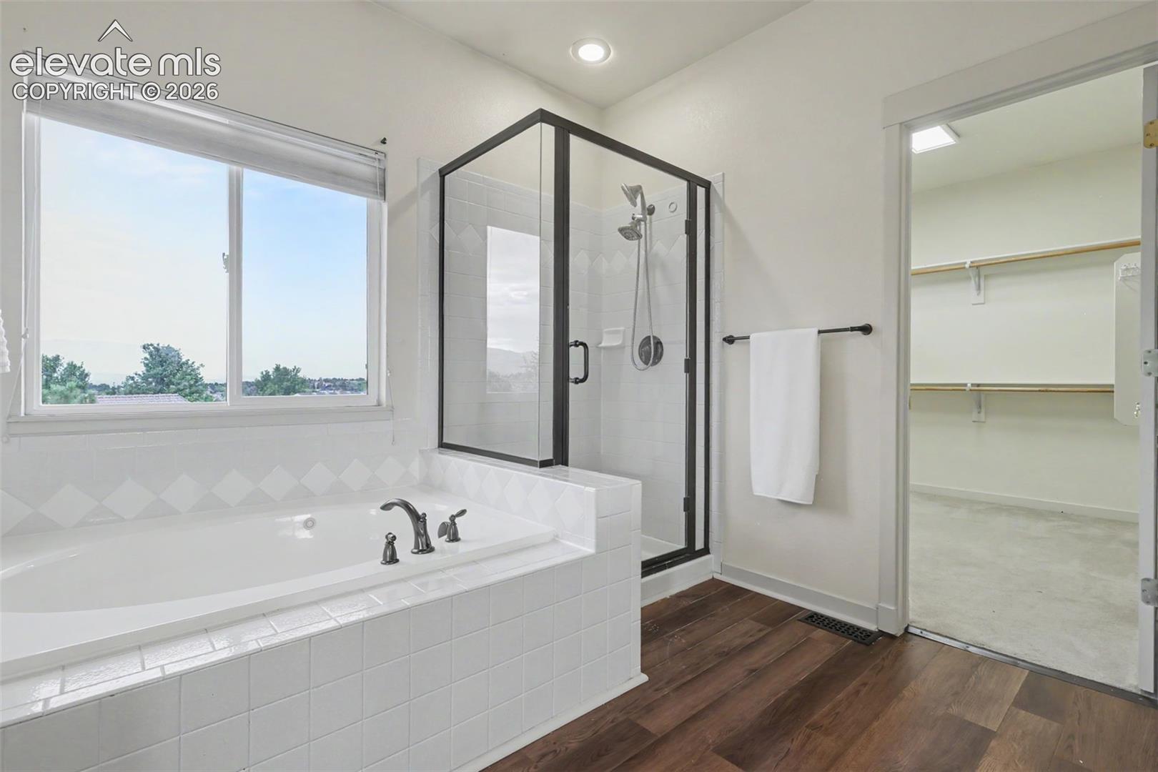 Bathroom featuring a built-in soaking tub with white tile surround, a glass-enclosed shower with black trim, and wood-finish flooring