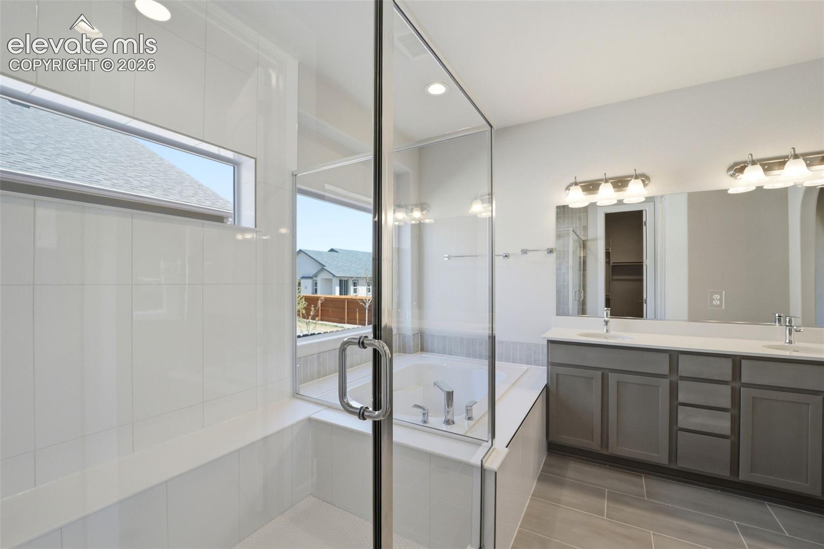 Bathroom featuring a frameless glass-enclosed shower with white wall tile, a built-in soaking tub, a dual-sink vanity with gray cabinetry, and large-format tile flooring