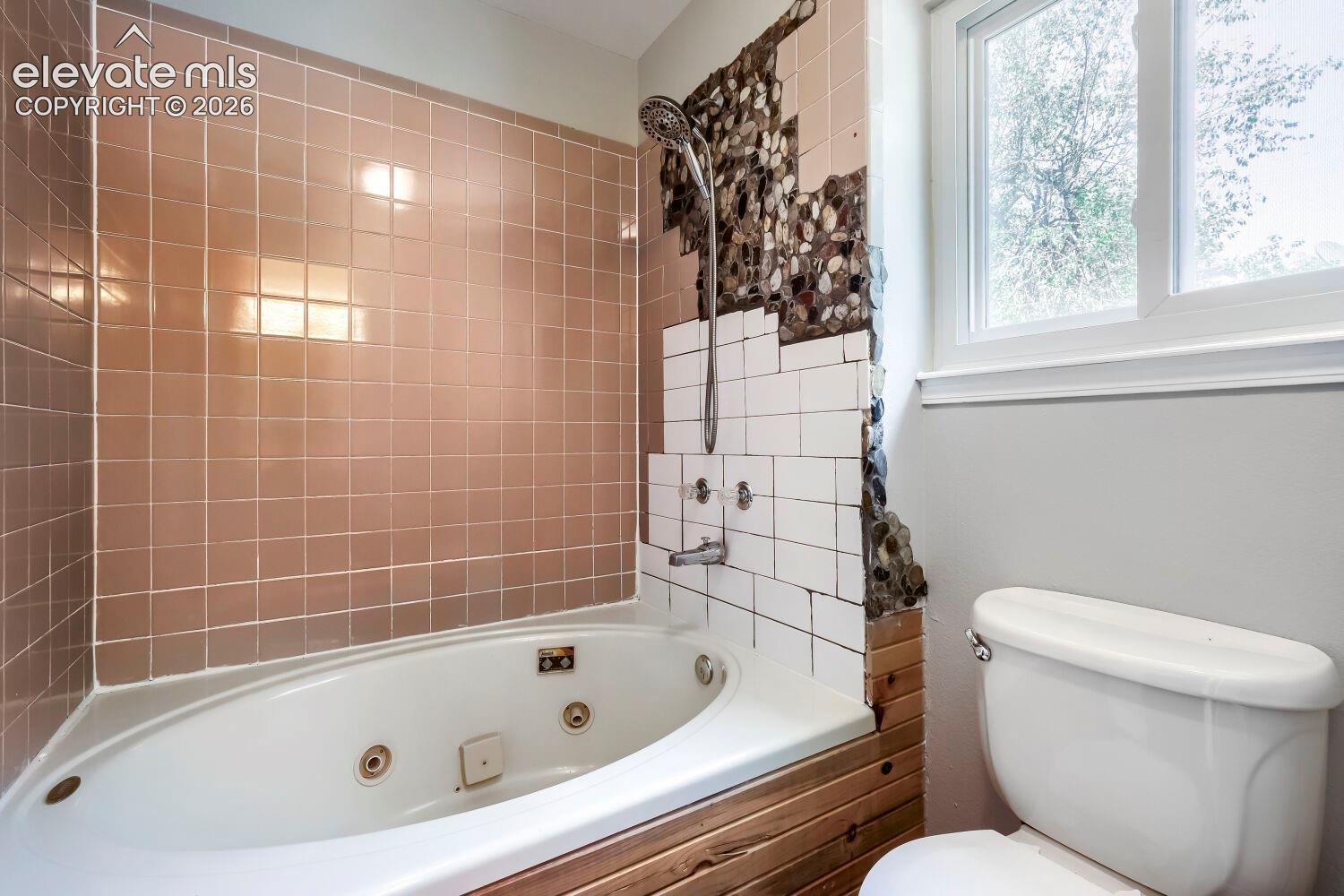 Jetted soaking tub with a tiled surround, complemented by a white toilet and a window