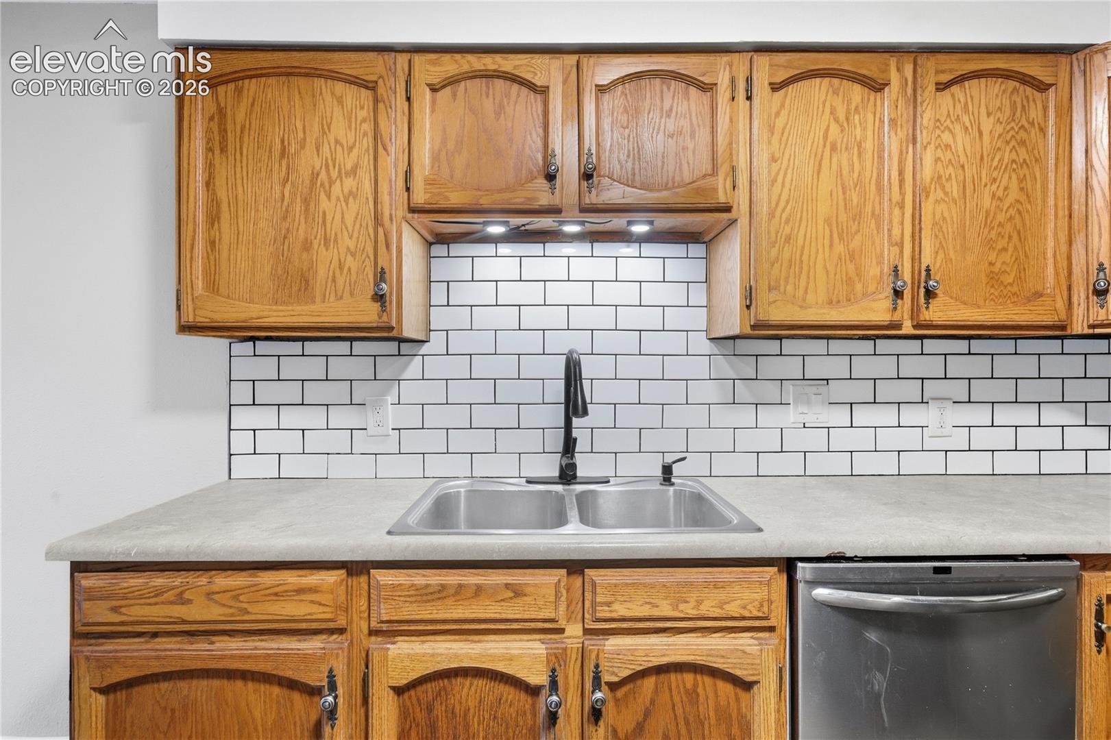 Wood cabinetry, dual basin stainless steel sink, subway tile backsplash, matte black faucet, and built-in dishwasher