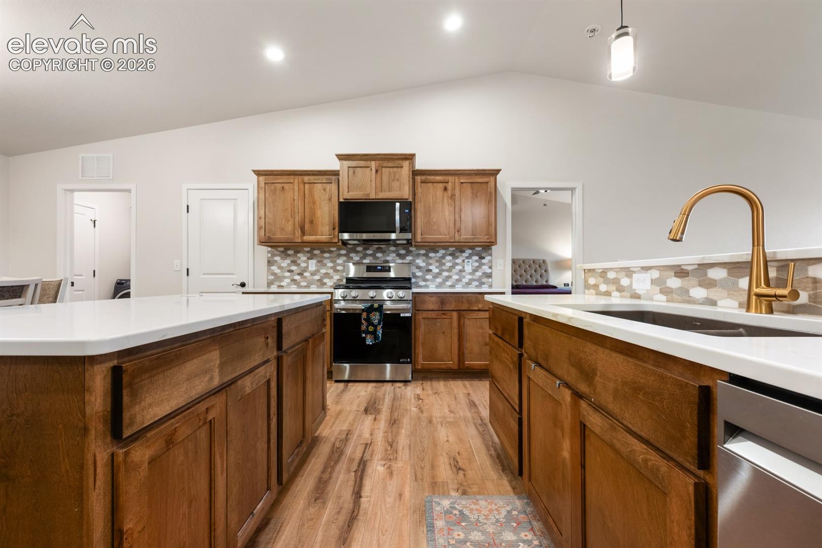 Kitchen featuring medium-tone wood cabinetry, white quartz countertops, stainless steel appliances, a hexagonal tile backsplash, and wood-finish flooring under a vaulted ceiling