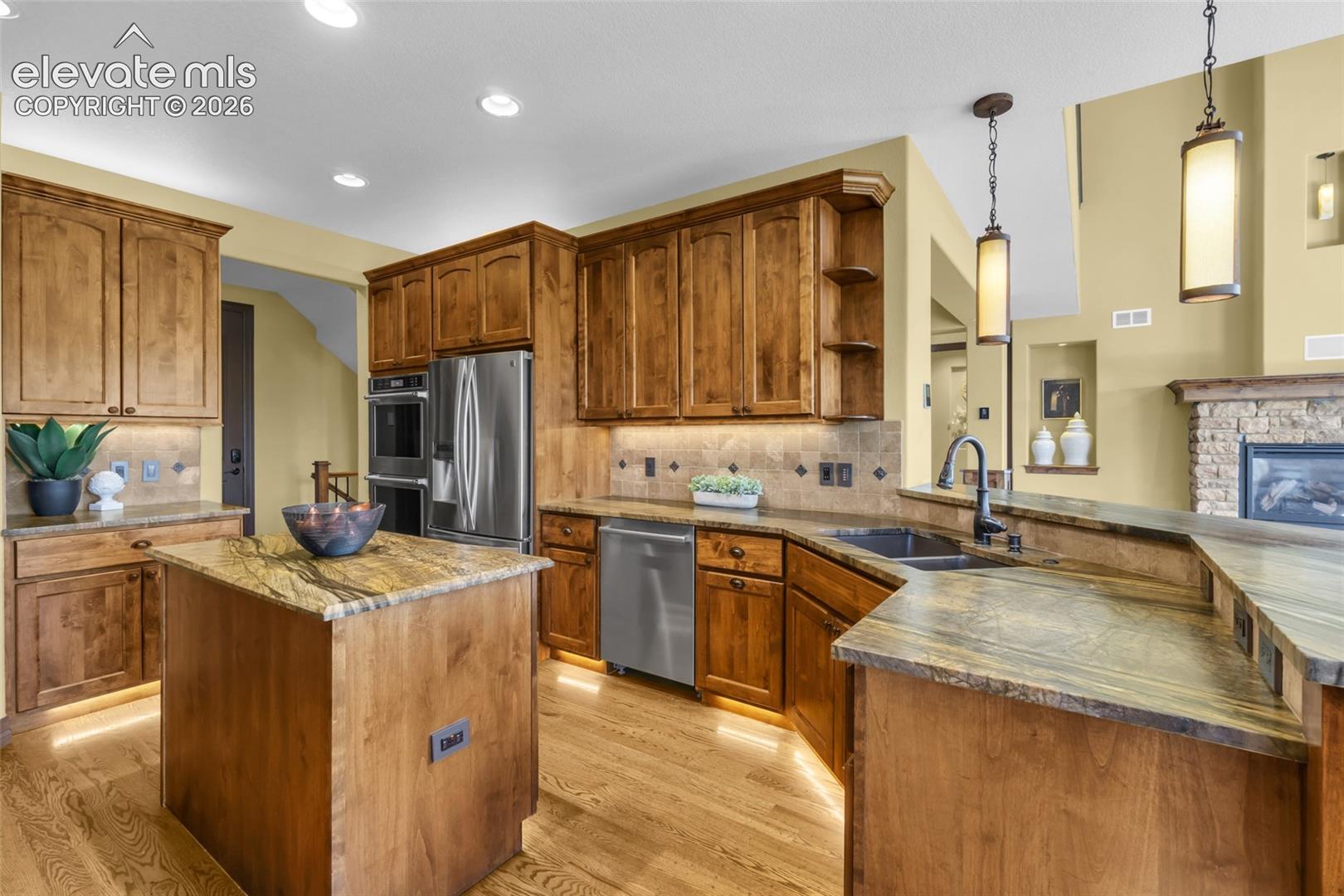 Kitchen featuring medium-tone wood cabinetry, stone countertops, stainless steel appliances, wood-finish flooring, and pendant lighting