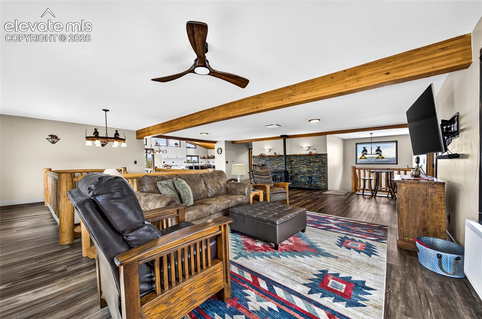 Open-concept living area featuring wood-finish flooring, exposed wood beams, a stone-clad fireplace, and a wood railing