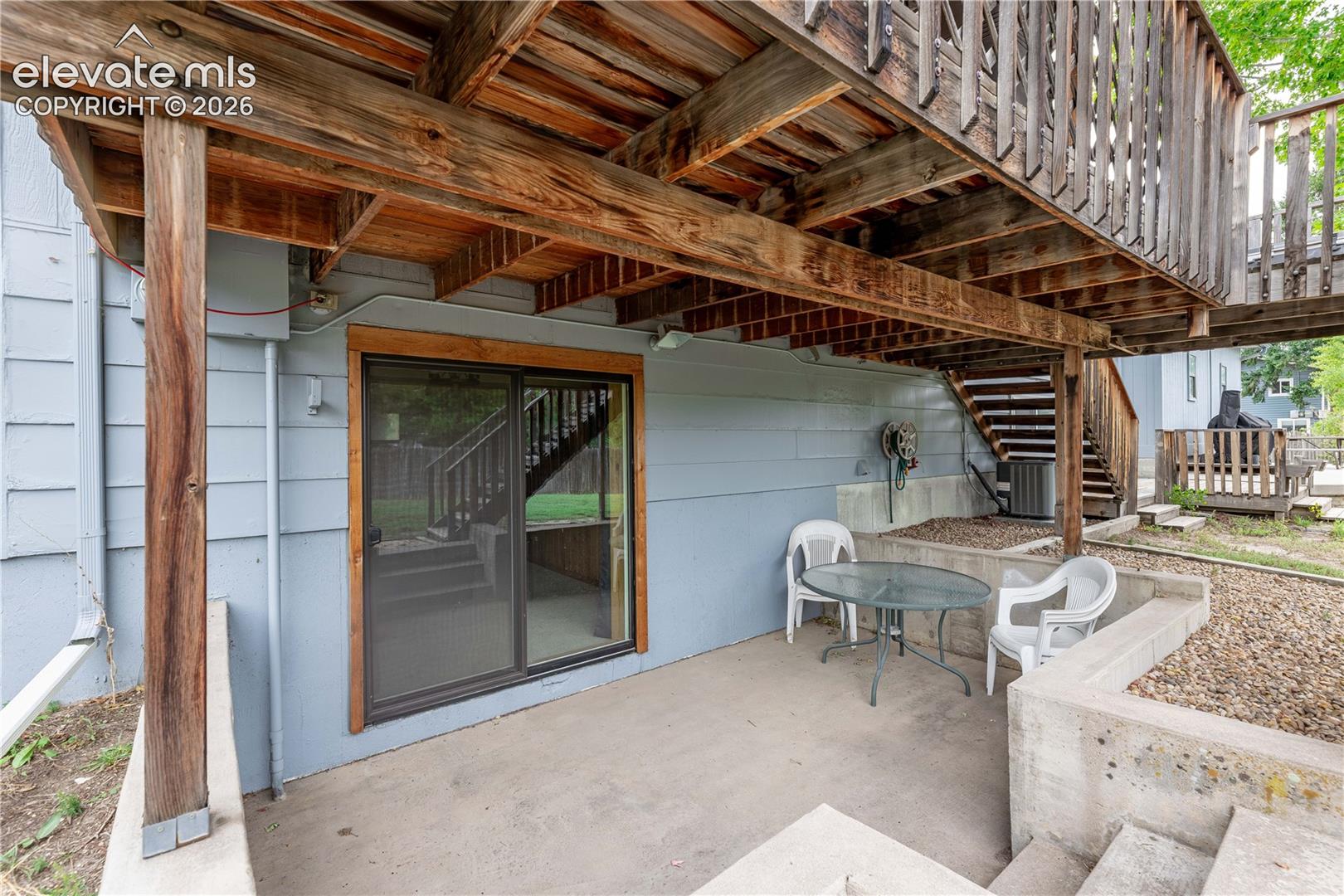 Concrete patio situated beneath an elevated wood deck featuring a sliding glass door and retaining wall elements