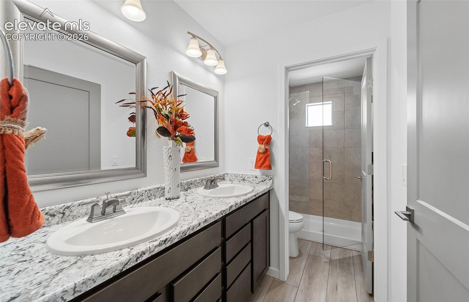 Dual-vanity cabinet with stone-finish countertop, undermount sinks, wood-finish flooring, and glass-enclosed shower with tile surround