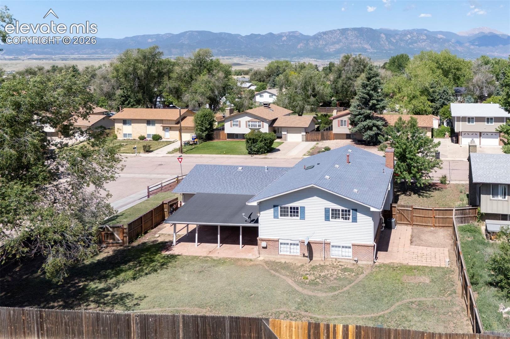 Aerial perspective showcasing a residential lot with a multi-level structure, covered patio, rear brick-paved area, enclosed wood fencing, and a mountain backdrop
