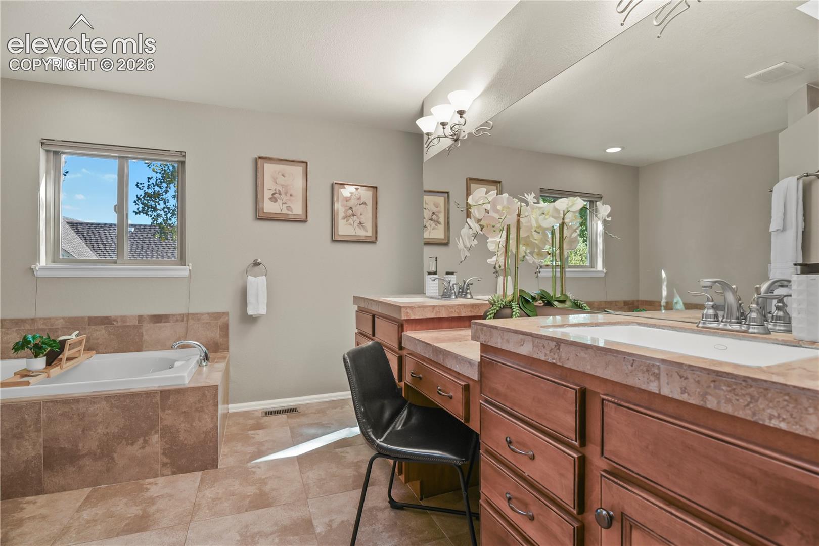 Dual-sink vanity with built-in make-up counter, wood-finish cabinetry, tile countertops, tile-surround soaking tub, and wall-to-wall mirror
