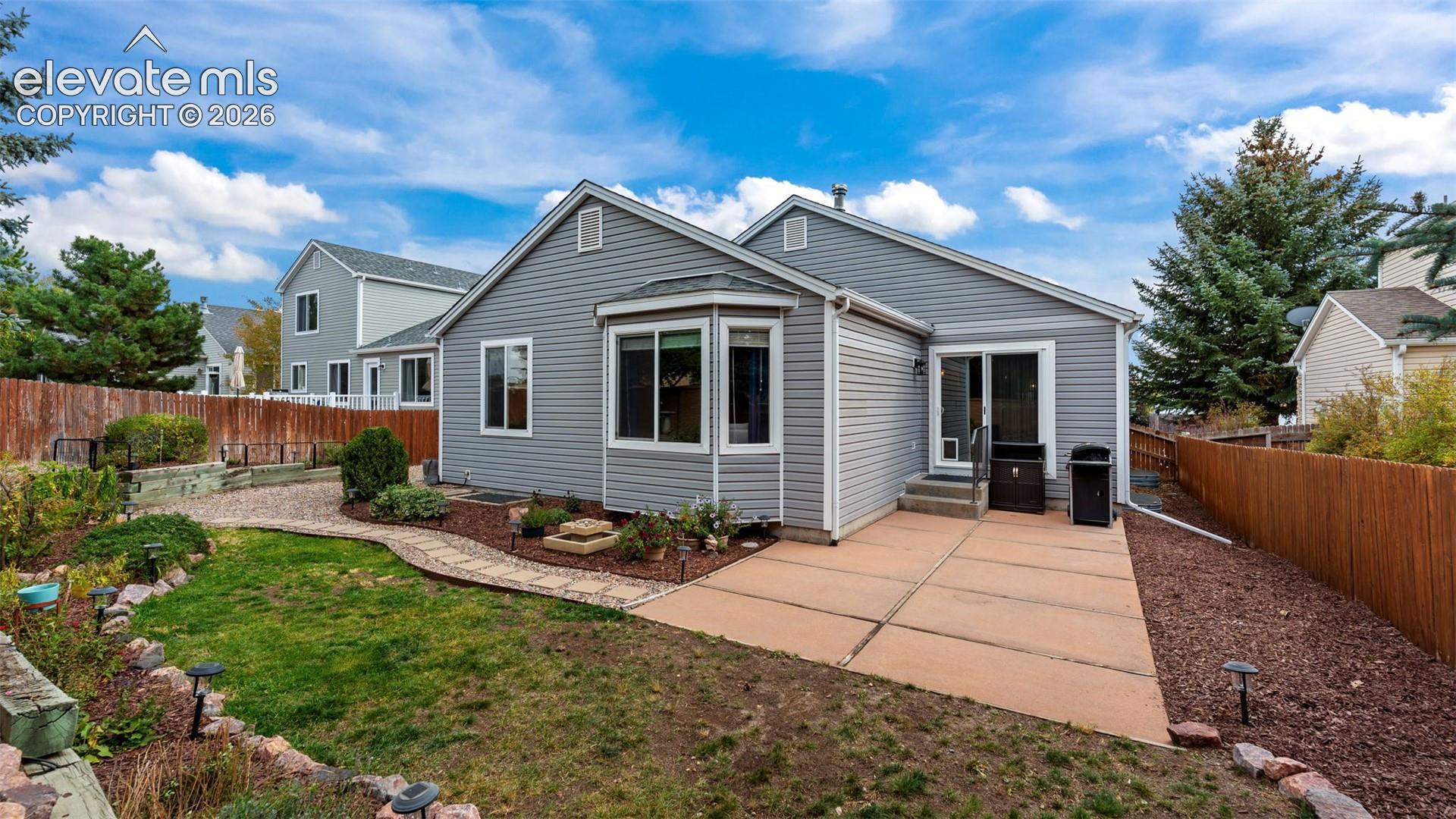 Exterior rear elevation featuring horizontal siding, a concrete patio, stone-bordered landscaping, and wood perimeter fencing