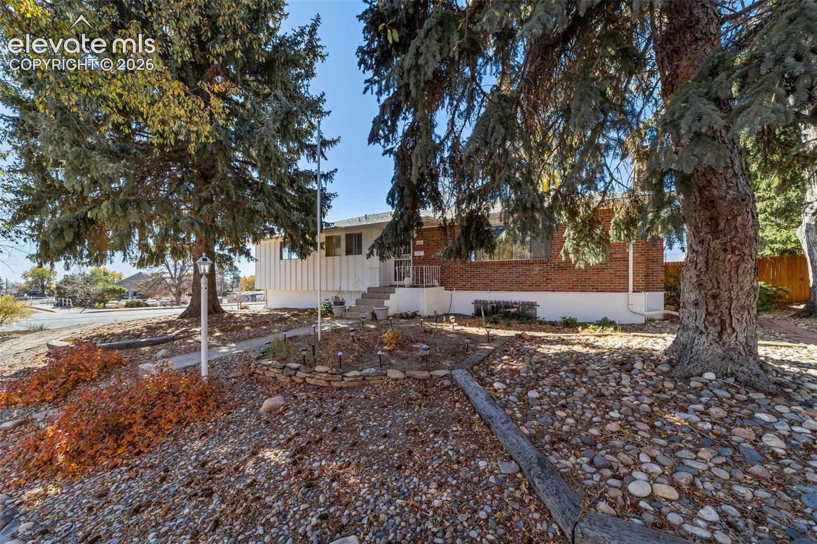Stone-landscaped front yard featuring mature evergreen trees, a brick facade, and a white sided exterior