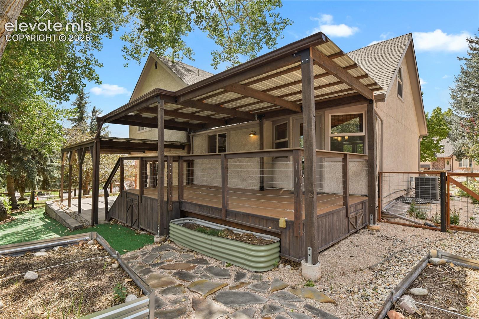 Stucco exterior featuring a covered composite deck with cable railing, metal roofing, integrated garden beds, and stone paver ground cover