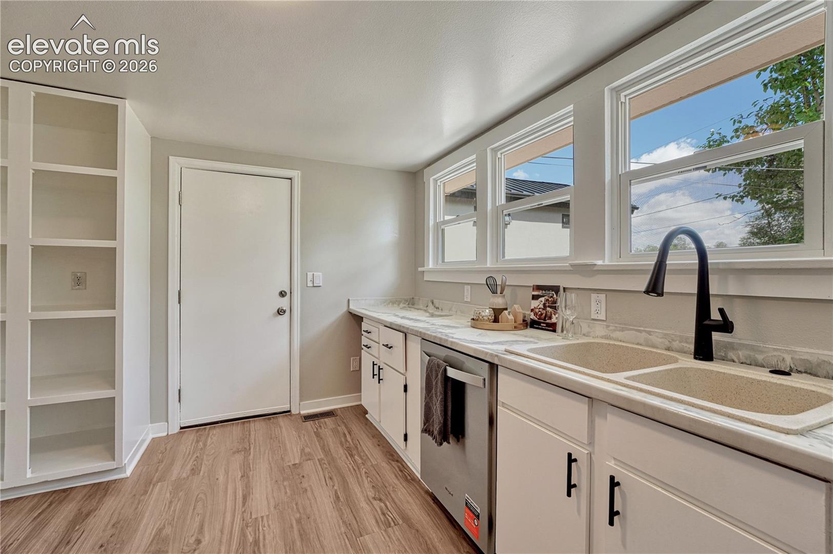 Modern kitchen featuring wood-finish flooring, white cabinetry with black hardware, a stainless steel dishwasher, a double basin sink with a black faucet, and three large windows