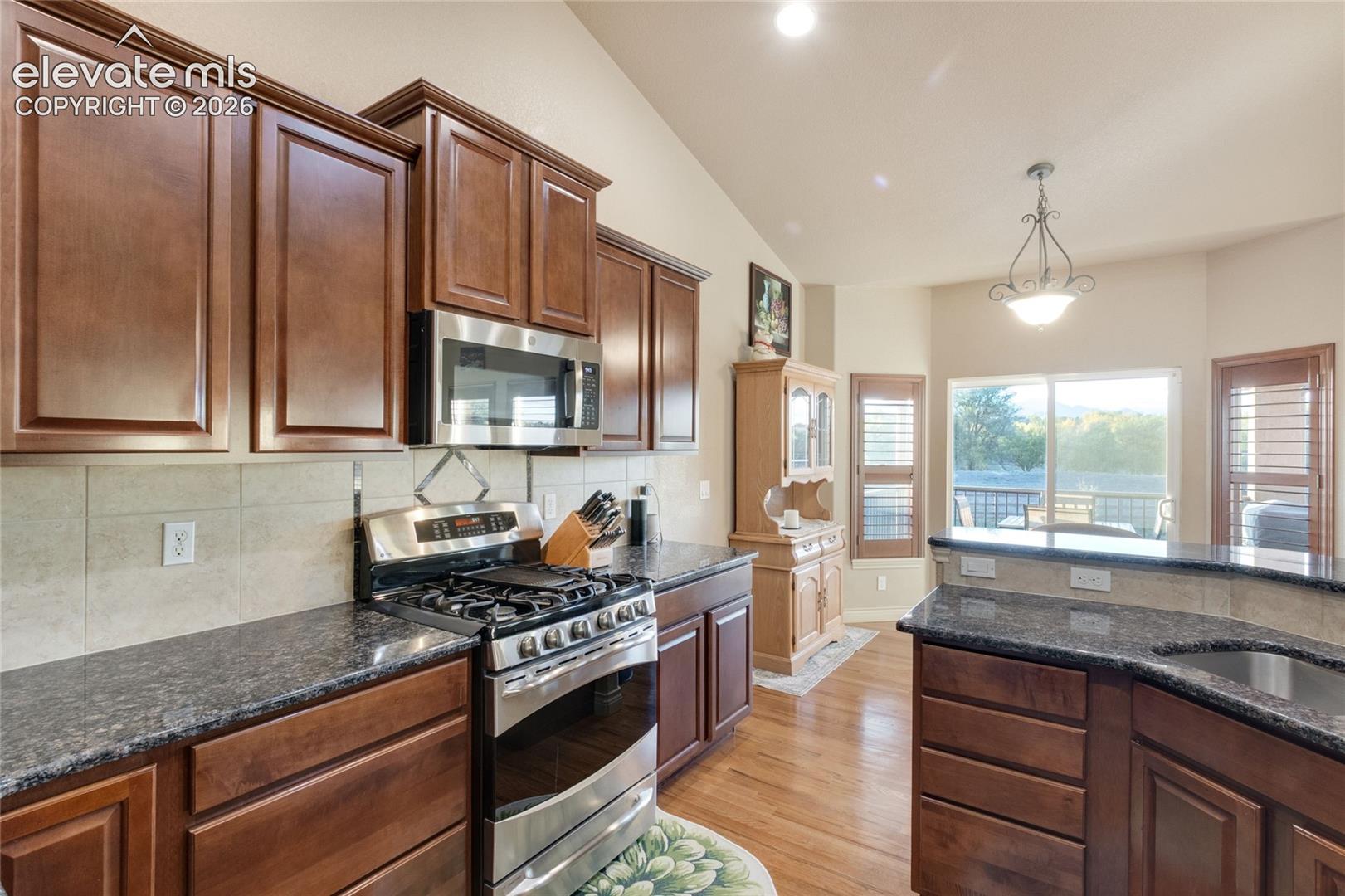 Kitchen featuring dark wood-finish cabinetry, stone countertops, stainless steel appliances, a tiled backsplash, and wood-finish flooring under a vaulted ceiling, breakfast area behind walks out to the deck