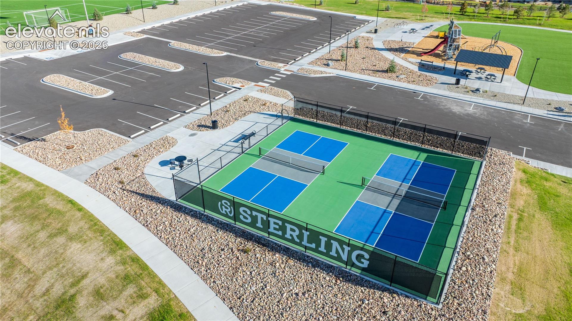 Community recreation area featuring dual pickleball courts, paved parking, athletic field, playground, and covered outdoor seating