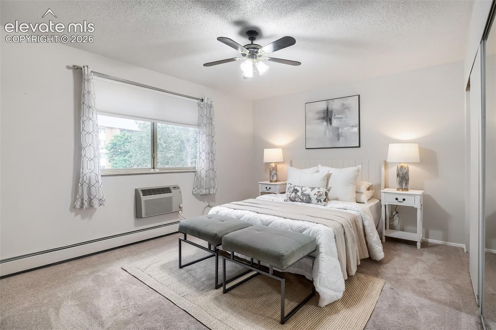 Carpeted room with a ceiling fan, two windows, baseboard heating, and an in-wall air conditioning unit