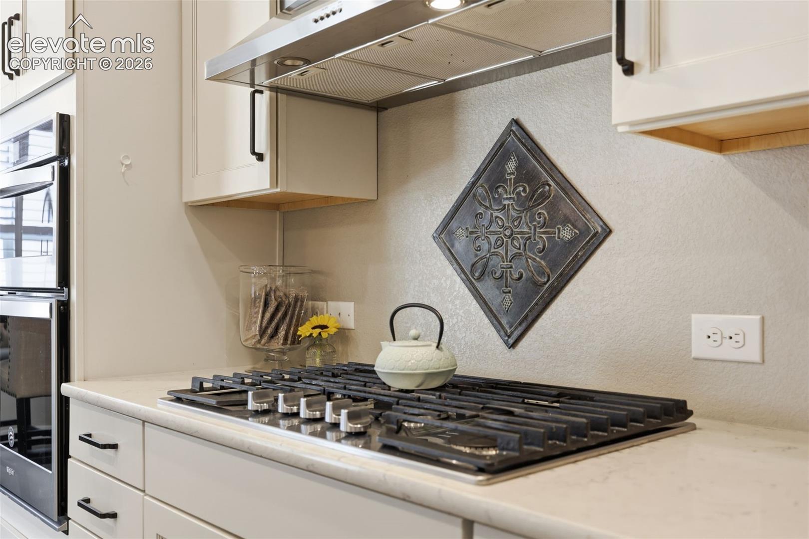 White shaker cabinetry with black hardware, stainless steal gas cook top, stainless steal hood vent and light color stone countertop.