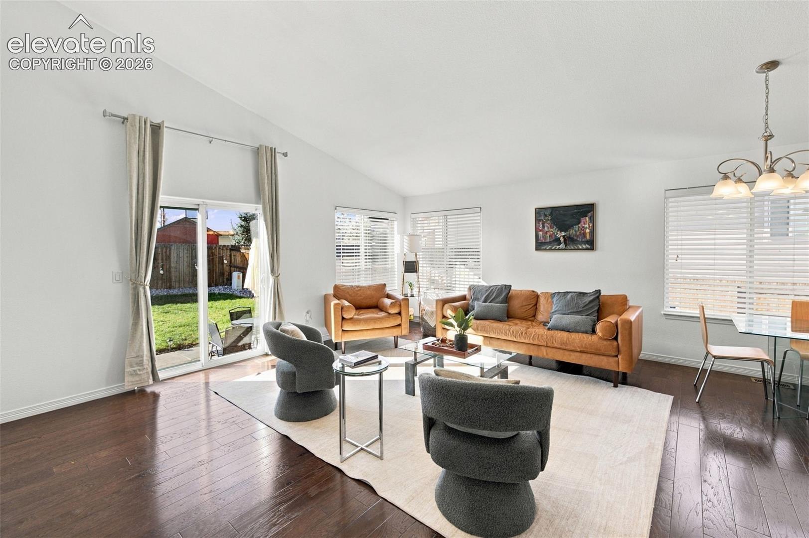 Vaulted ceilings, wood-finish flooring, sliding glass door, multi-pane windows with horizontal blinds, and a multi-arm chandelier