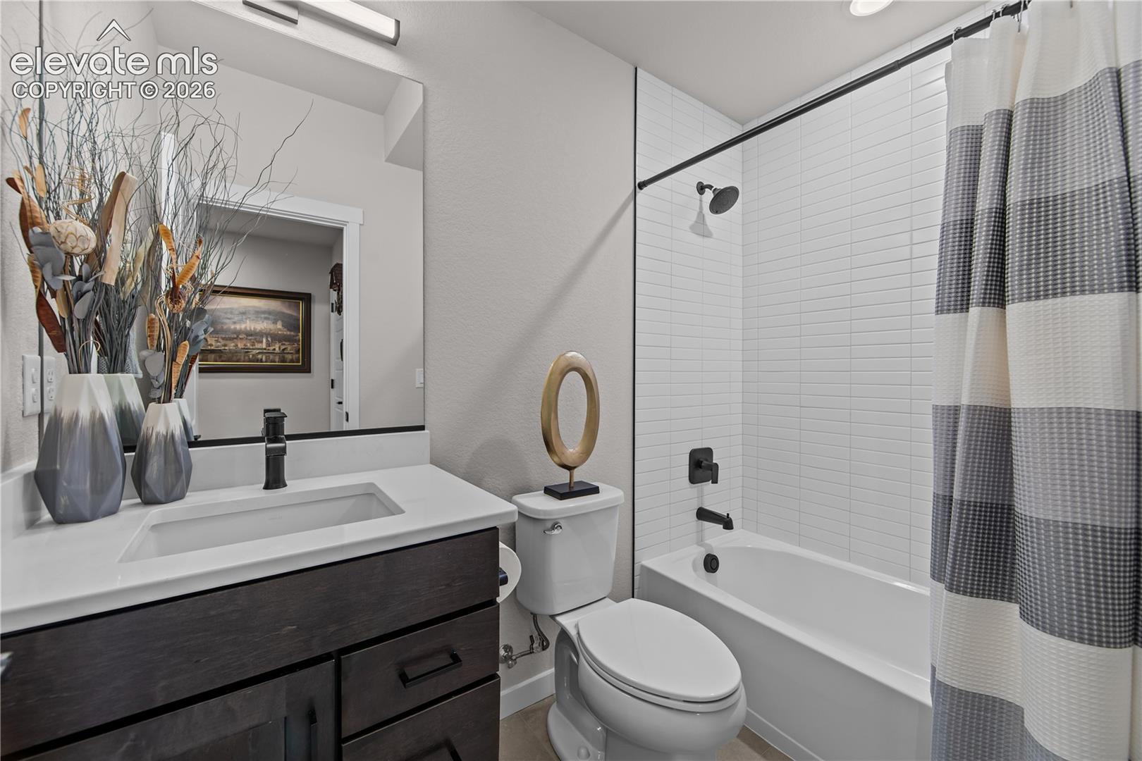 Dark-toned wood vanity with a white countertop and rectangular basin, paired with matte black hardware, a large wall-mounted mirror, and a tub-shower combination featuring stacked white wall tile