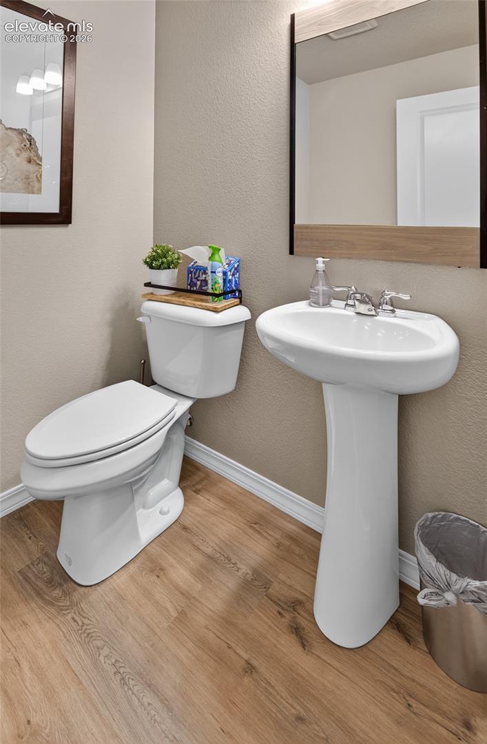 White pedestal sink with a dual-handle faucet, elongated toilet, and rectangular mirror featuring a wood-finish frame over wood-finish flooring