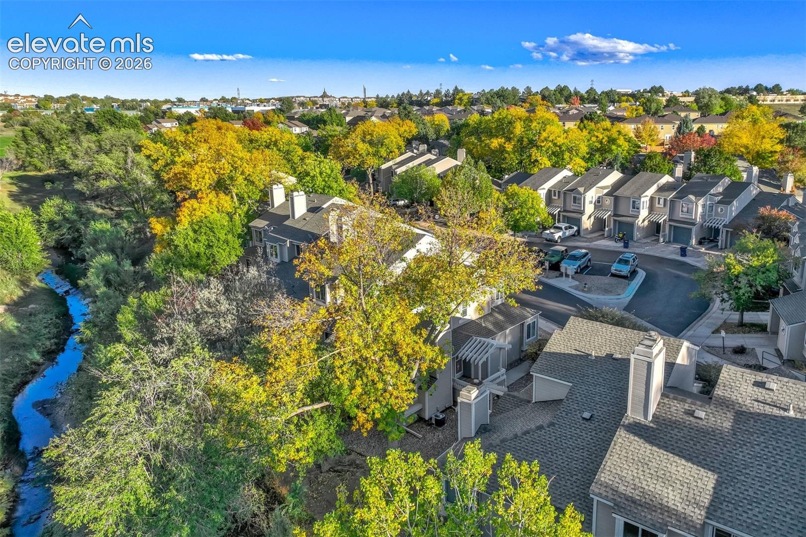 Aerial perspective of a multi-unit residential complex featuring mature deciduous trees, a winding natural creek, paved cul-de-sac streets, and attached pergolas