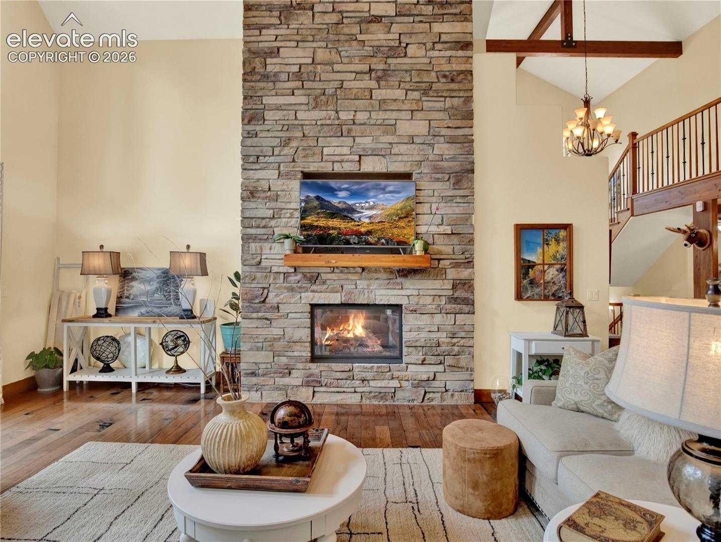 Stone fireplace, to the left is the floor to ceiling windows with doors to the outdoor front deck. To the right is open kitchen with loft above