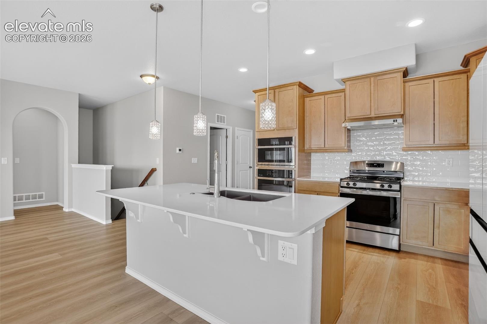 Open-concept kitchen featuring a spacious island with a built-in sink, stainless steel appliances, light wood cabinetry, white subway tile backsplash, and light wood-finish flooring