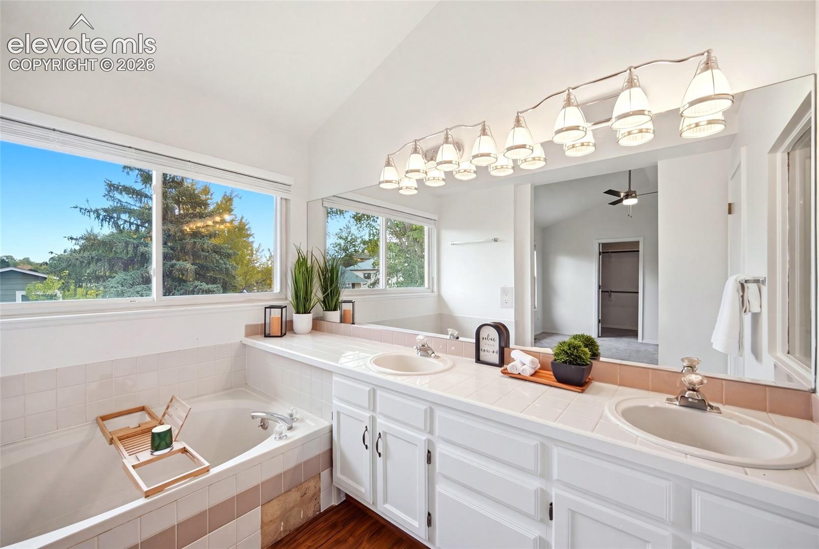 Double vanity with tile countertops, integrated dual sinks, large wall-to-wall mirror, multi-light fixture, and built-in soaking tub with tile surround