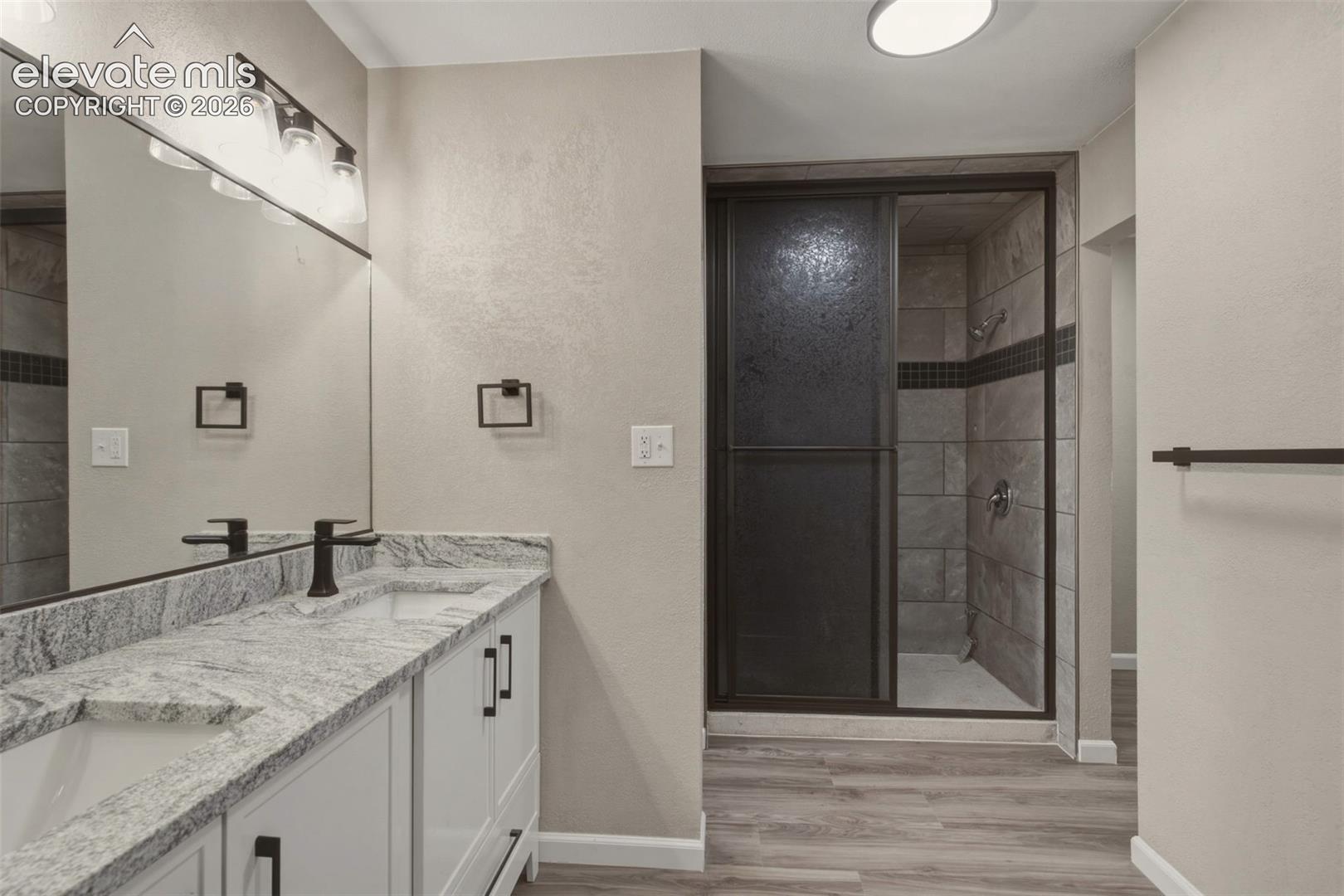 Dual vanity with granite countertop, white cabinetry, matte black fixtures, wood-finish flooring, and a tiled shower with dark-framed glass enclosure