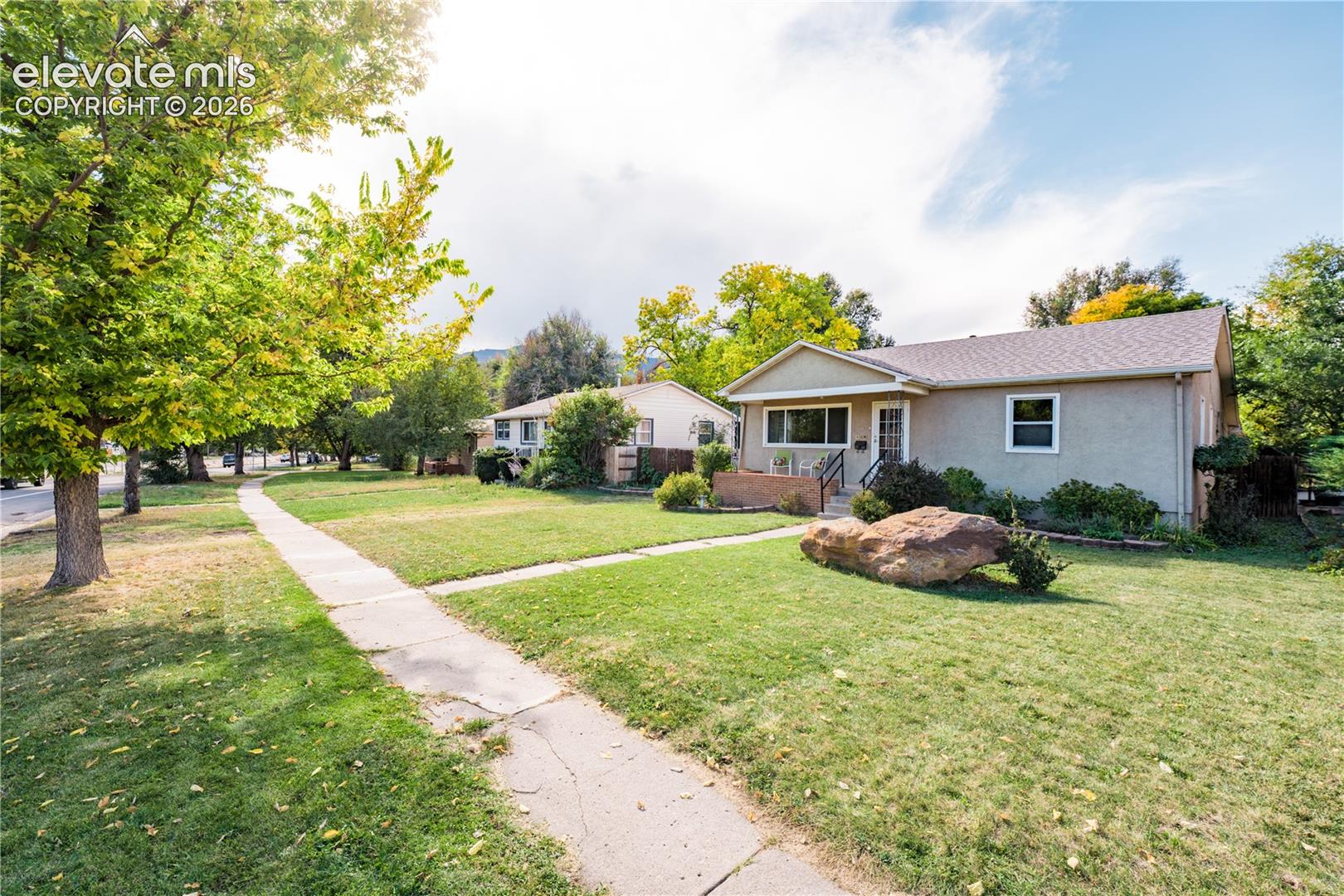 Stucco exterior with a pitched roof, featuring a covered porch with dark railings, a well-maintained lawn, and established trees