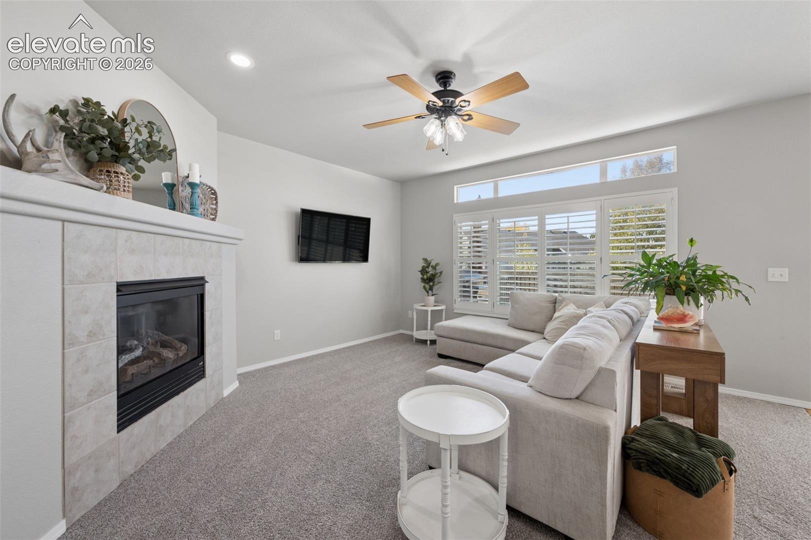 Living area featuring a fireplace with a neutral tile surround, custom transom window, plantation shutters, ceiling fan with light fixture, and recessed lighting
